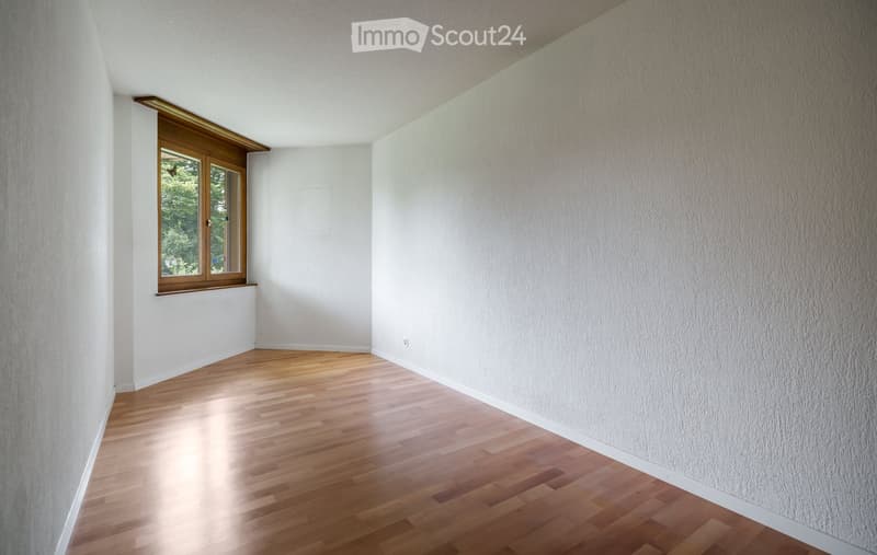Empty room with wooden flooring, white walls, and a window with wooden frame showing greenery outside.