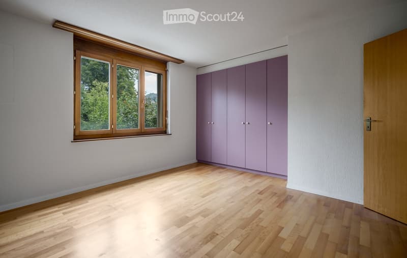 Empty room with light wooden flooring, white walls, large window with wooden frame, and built-in purple cabinets.