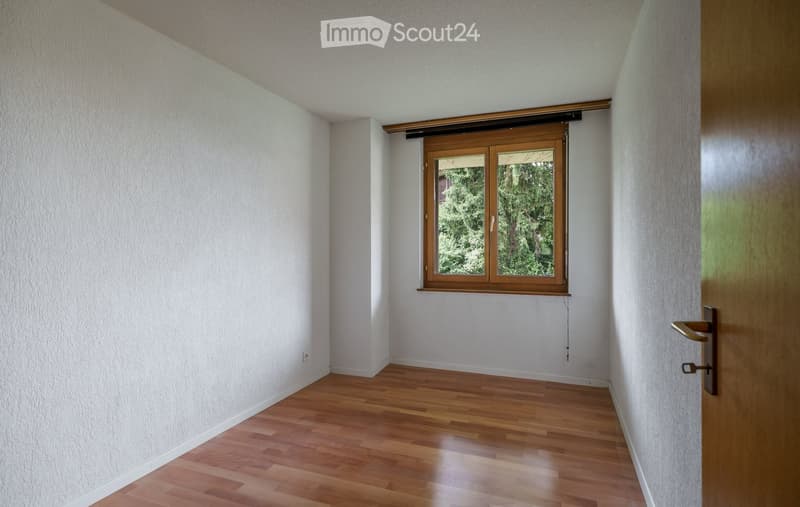 Empty room with wooden flooring, white walls, and a window with wooden frame showing greenery outside.