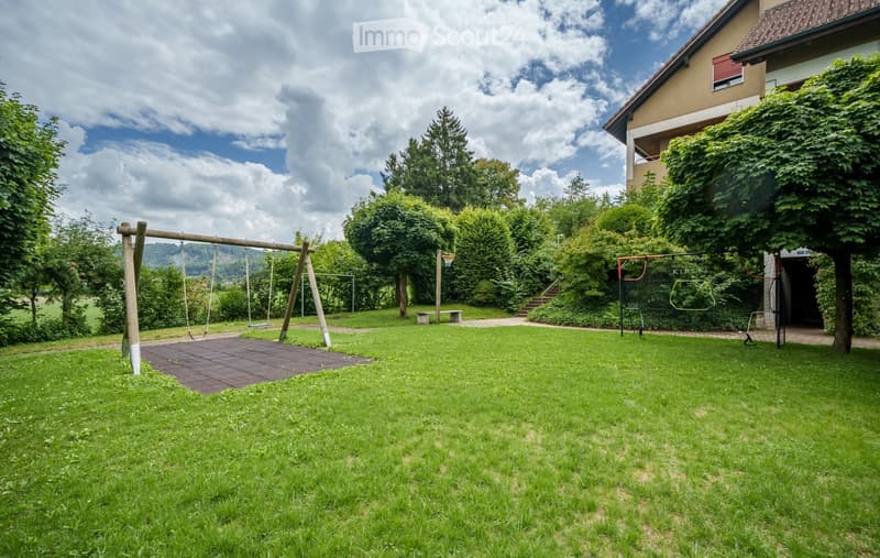 Lush green garden with a playground featuring swings and a climbing frame, surrounded by trees and a two-story house with a balcony.