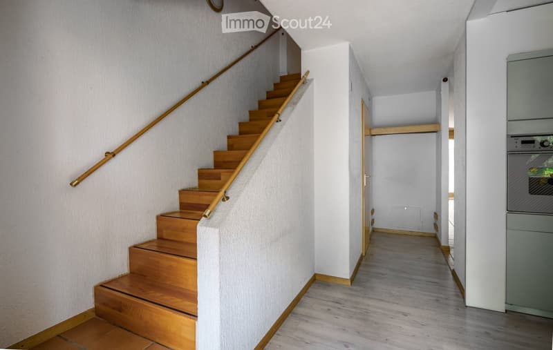 A modern hallway with a wooden staircase featuring a metal railing, light-colored walls, and a grey appliance on the right side.