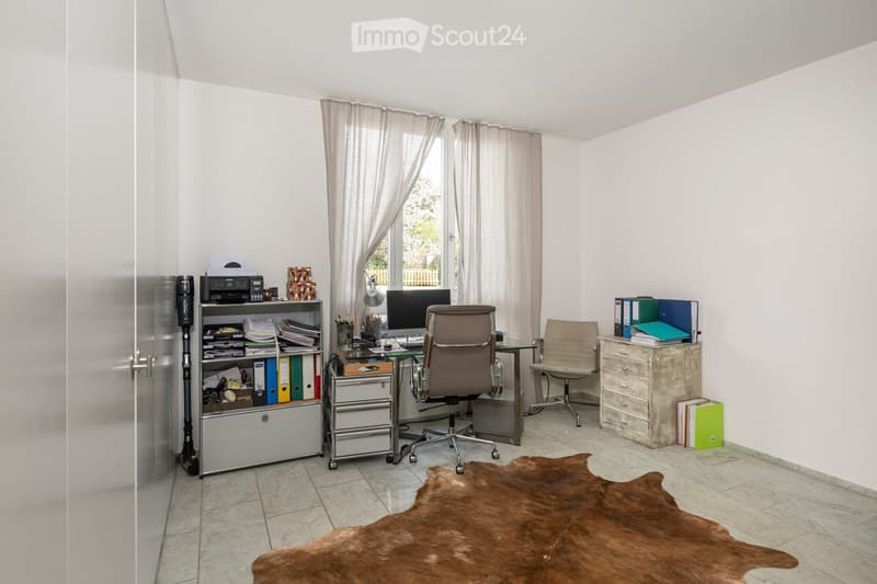 Bright office space with white walls, tiled floor, large window with curtains, office chair, desk with computer monitor, printer, bookshelf, and a cowhide rug on the floor.