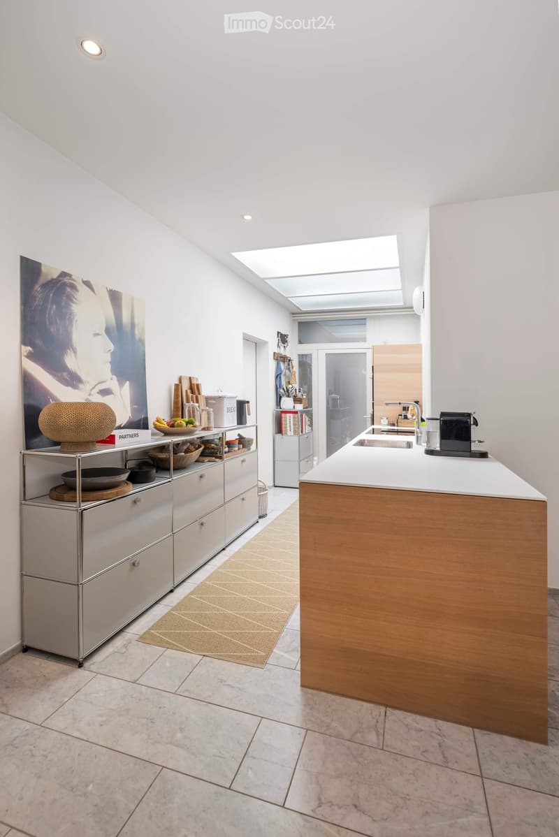 Modern kitchen with white walls, marble flooring, and a central island with wooden accents. Features include a stainless steel cabinet, a sink with a modern faucet, a coffee maker, and a bowl of fruit on the counter.