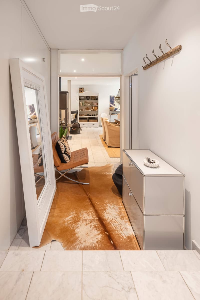 Modern hallway with a large mirror, a sleek white cabinet, a brown leather chair, and a small table with a decorative item on it. The floor is polished wood, and the walls are white.