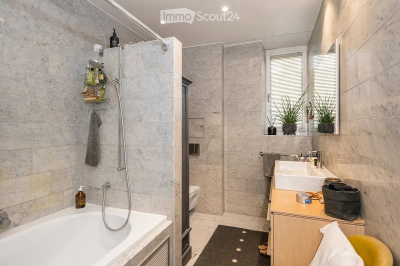 Modern bathroom with marble tiles, a bathtub, a shower, a double sink, a large mirror, and decorative plants on the windowsill.