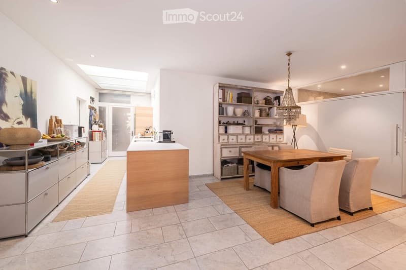 Modern kitchen with white cabinets, a central wooden island, a dining table with four chairs, and built-in shelving. The space features a chandelier, a refrigerator, and a sink area with a coffee machine.