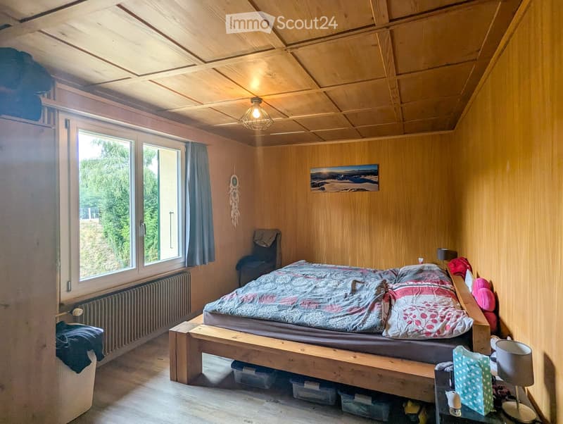 Cozy bedroom with wooden walls and ceiling, large window with view, bed with colorful bedding, chair, and decorative items.