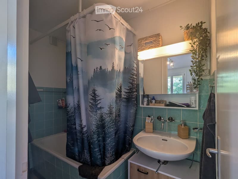Bathroom with a bathtub, shower curtain, sink, mirror, and various toiletries. Green tiles and wooden cabinetry.
