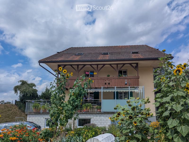 Two-story house with a tiled roof, wooden balcony, and sunflowers in the garden.