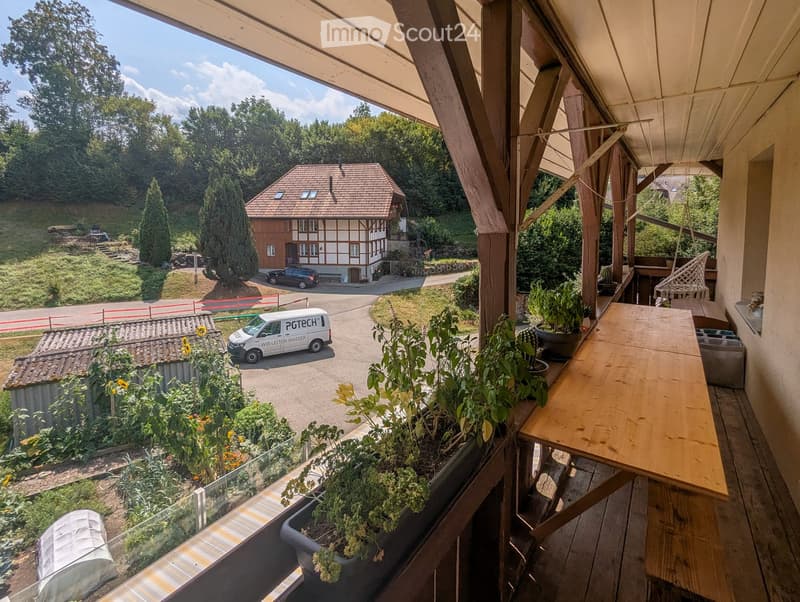 Balcony with wooden table, bench, and potted plants, overlooking a driveway and a traditional house.
