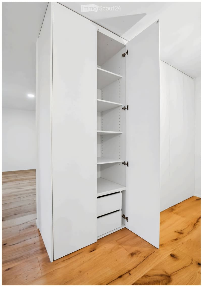 An empty room with white walls and wooden flooring, featuring a built-in white cabinet with open shelves and drawers.