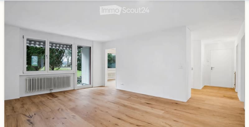 Empty room with light wooden flooring, white walls, large windows, and a radiator. No furniture or equipment present.