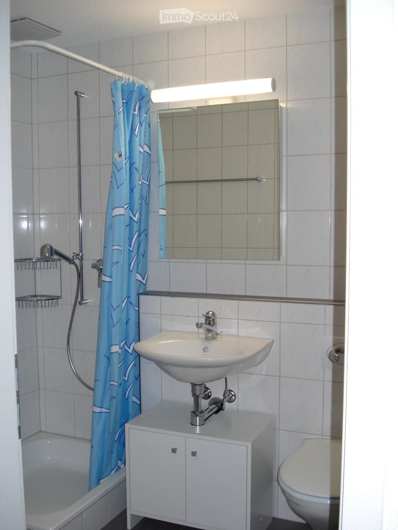 Modern bathroom with white tiles, a sink with a chrome faucet, a mirror above it, a bathtub with a blue shower curtain, a toilet, and a small cabinet under the sink.
