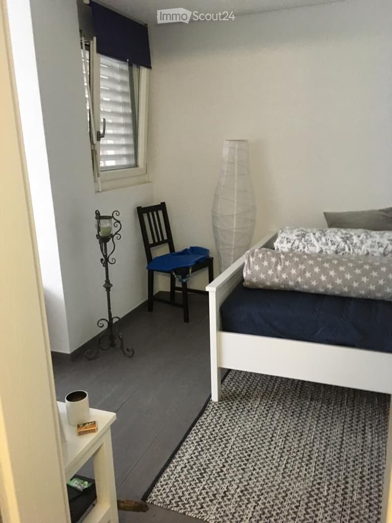 Cozy bedroom with a white bed, dark blue bedding, a patterned rug, a chair with a blue cushion, a small table with a mug, and a decorative lamp near the window.