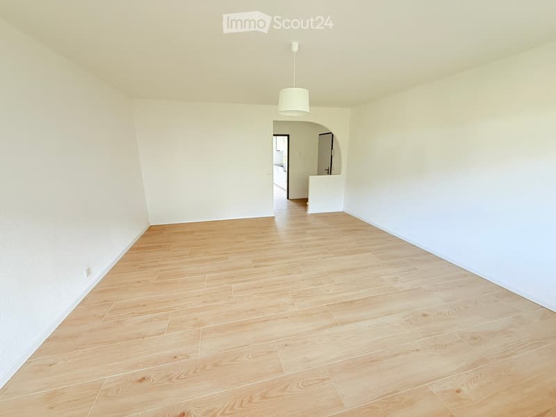 Empty room with light wooden flooring and white walls, featuring a hanging light fixture and an open doorway leading to another room.