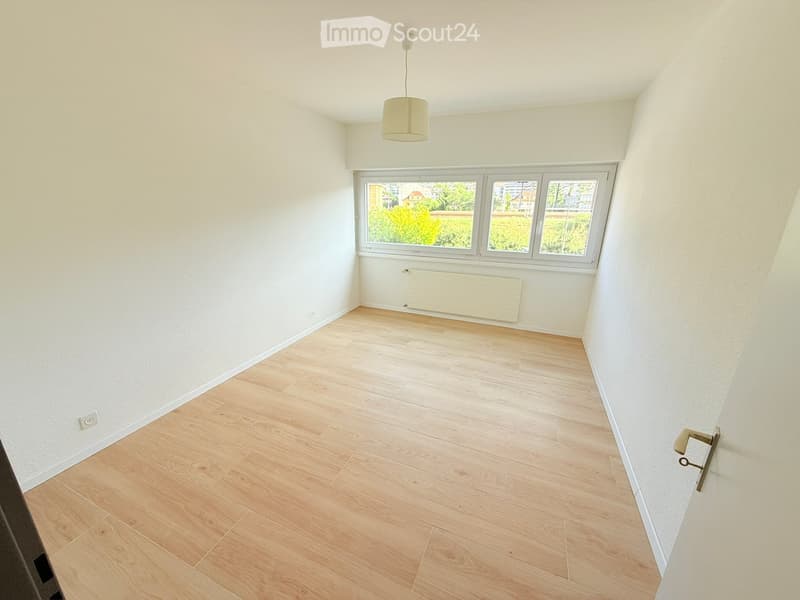 Empty room with light wooden flooring, white walls, large window with view, and a hanging light fixture.