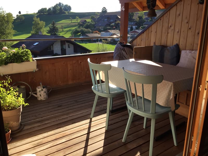 Wooden balcony with a table, chairs, and a small sofa, overlooking a scenic countryside view.