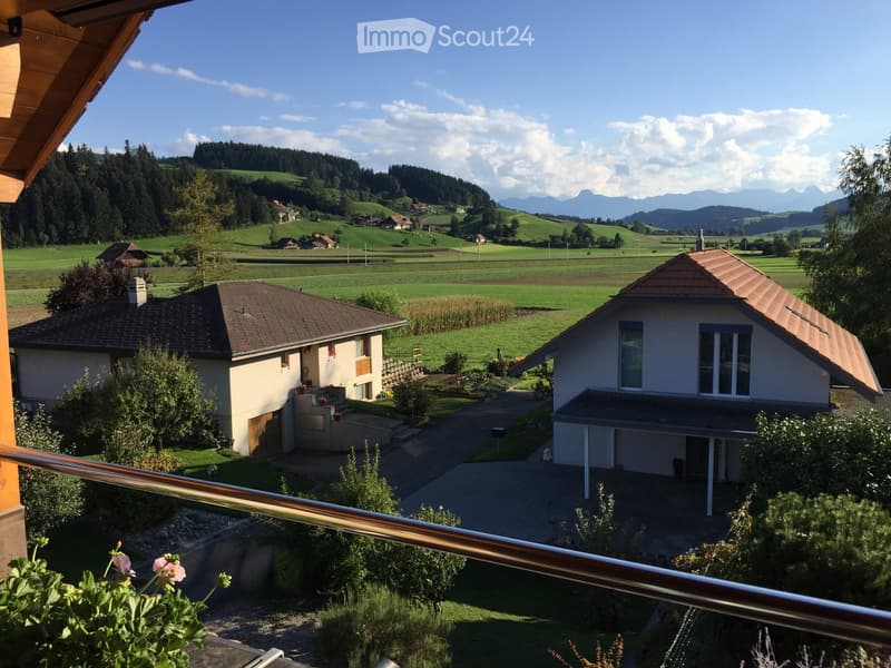 View from a balcony overlooking a rural landscape with houses, green fields, and distant mountains.