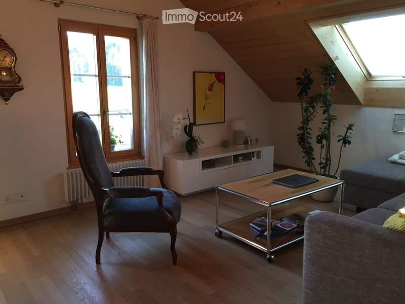 Cozy living room with wooden floors, large windows, a sofa, armchair, coffee table with laptop and books, a table lamp, and decorative plants.