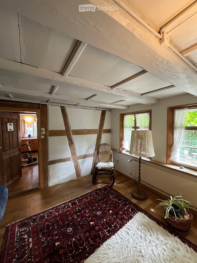 Cozy attic room with wooden floors, exposed wooden beams, large windows with white curtains, a patterned rug, a wicker chair, a floor lamp, and a small plant in a basket. An exercise bike is visible in the adjacent room.