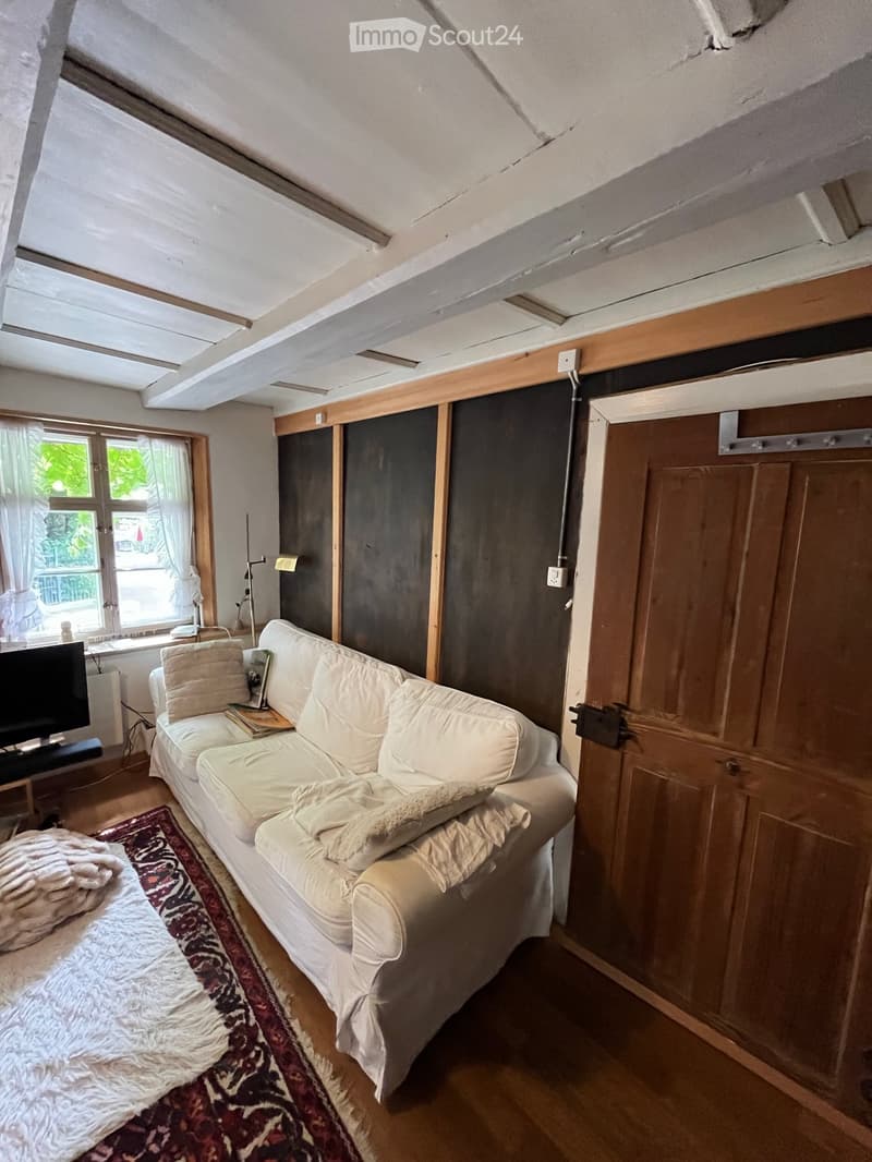 A cozy living room with a white sofa, pillows, a blanket, a small table with books, and a television. The room features wooden walls, a large window with curtains, and a patterned rug.