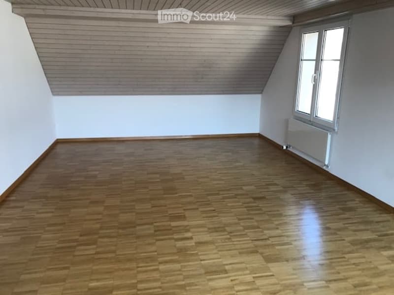 Empty room with wooden flooring and white walls, featuring a slanted ceiling and a single window with a radiator below it.