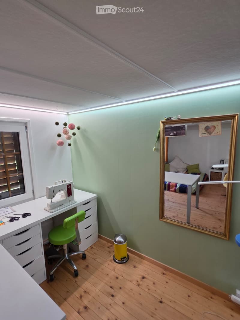 A small, well-lit room with a sewing machine on a desk, a green chair, a large mirror, and a trash can. The room has wooden flooring and light green walls.