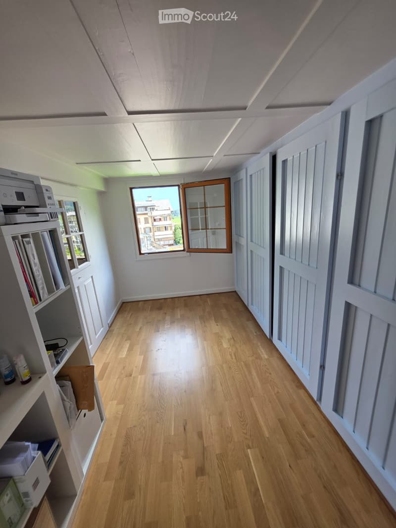 Empty hallway with wooden flooring, white walls, and several doors. A window at the end provides natural light, and a shelf with books and a printer is visible on the left side.
