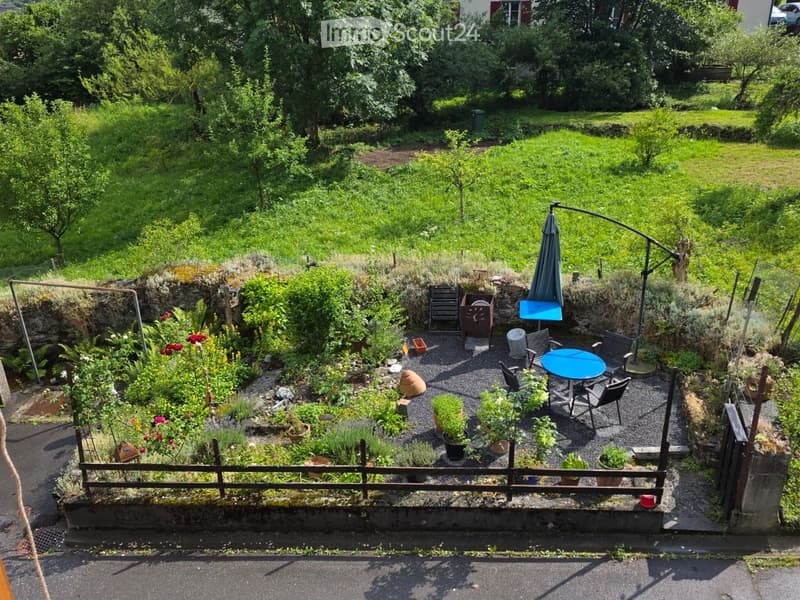 A small, well-maintained garden with a seating area featuring a blue table, chairs, and an umbrella. The garden includes various potted plants and is enclosed by a wooden fence.