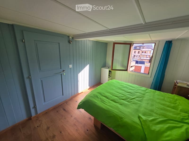 Cozy bedroom with wooden flooring, green bedspread, nightstand, and window with blue curtains.