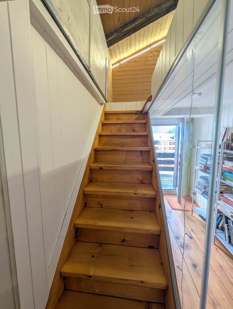 Wooden staircase with white walls and a glass railing, leading to a room with bookshelves and a view of the outside.