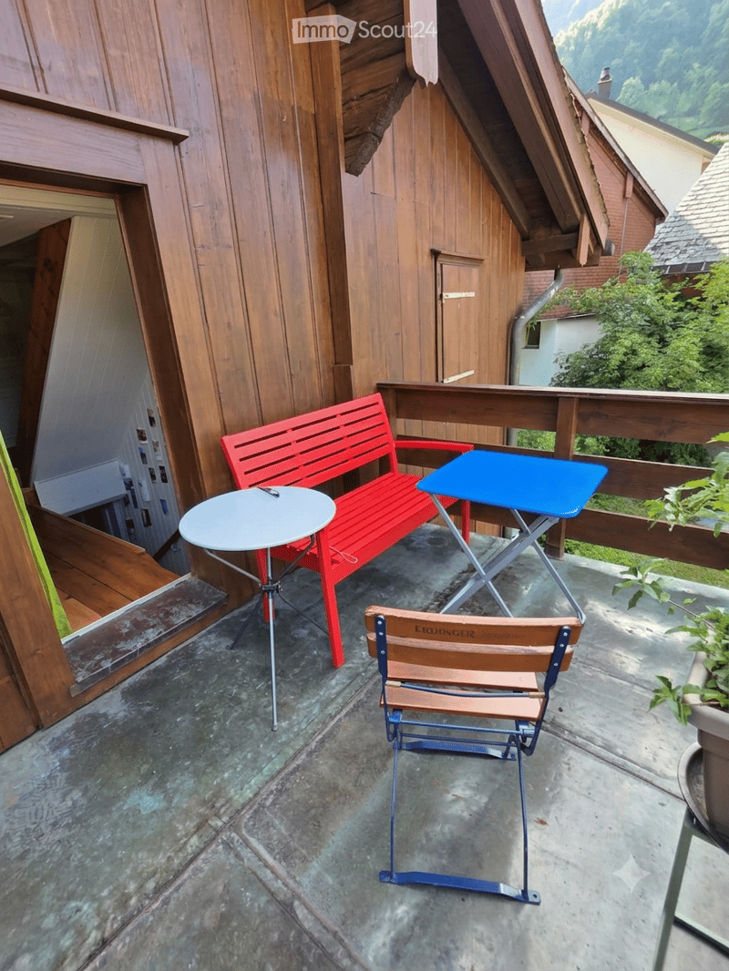Balcony with a red bench, white round table, blue folding chair, and scenic mountain view.