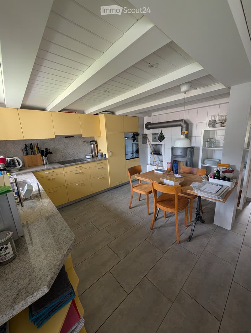 Bright kitchen with yellow cabinets, a central dining table with chairs, and various appliances including an oven, fridge, and dishwasher. The space features a modern design with a slanted ceiling and ample natural light.