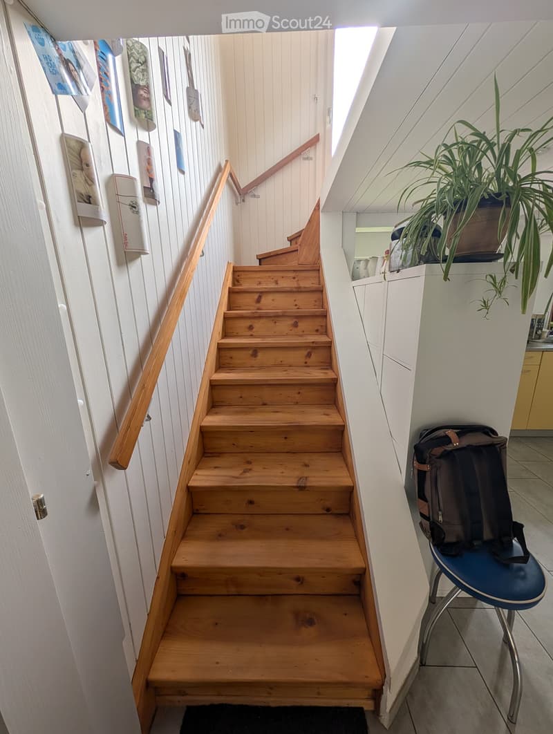Wooden staircase with a handrail, white walls, and posters on the wall. A blue chair with a backpack is placed near the staircase, and a potted plant is visible in the background.