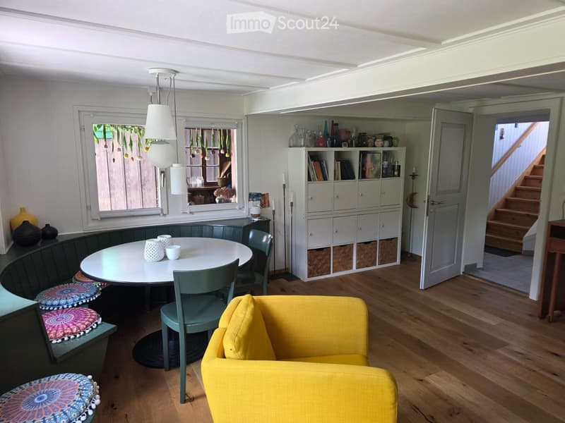 Bright living and dining area with wooden flooring, a yellow sofa, a round dining table with four chairs, colorful stools, a built-in bookshelf, and decorative vases.