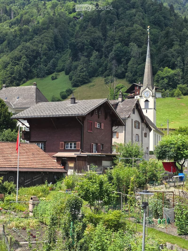 A traditional Swiss house with a steeply pitched roof, surrounded by lush greenery and a small garden area. A nearby church with a tall spire is visible in the background, set against a backdrop of green hills and trees.
