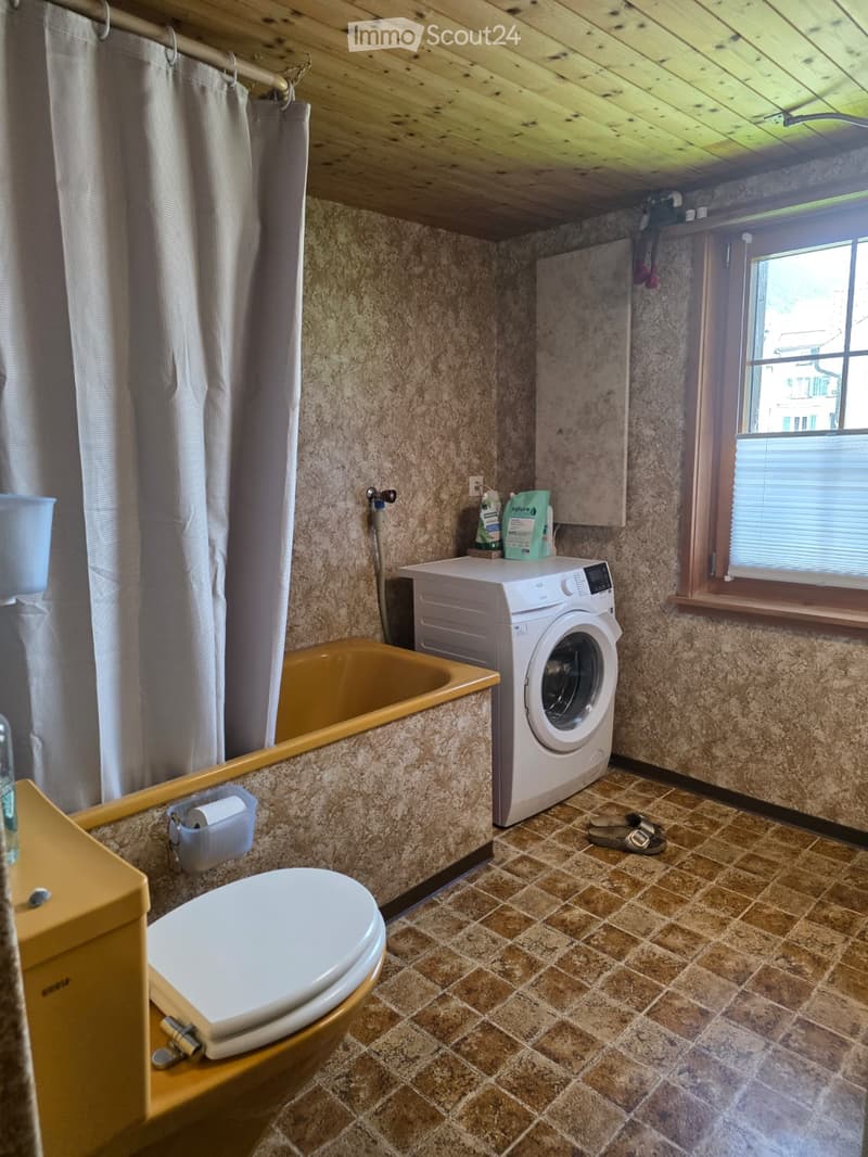 Bathroom with a toilet, bathtub, washing machine, and shower curtain. The room features tiled walls and a window with blinds.