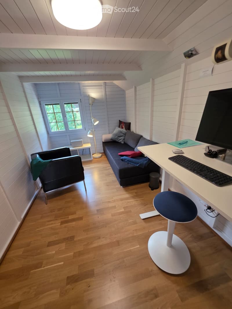 A compact, multi-functional room with a sofa, desk, and chair, featuring wooden flooring and white walls.