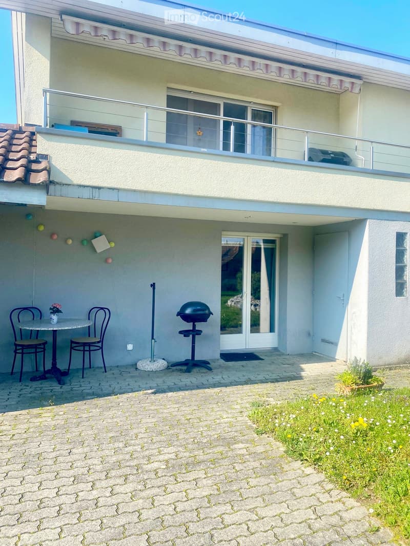 Outdoor patio area with a small table, two chairs, and a barbecue, adjacent to a modern house with a balcony and glass railing.