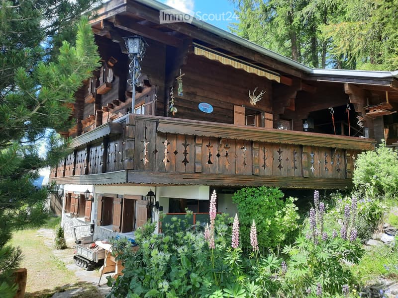 A two-story wooden house with a balcony, intricate wooden railings, and a stone base. The house is surrounded by lush greenery and flowers.