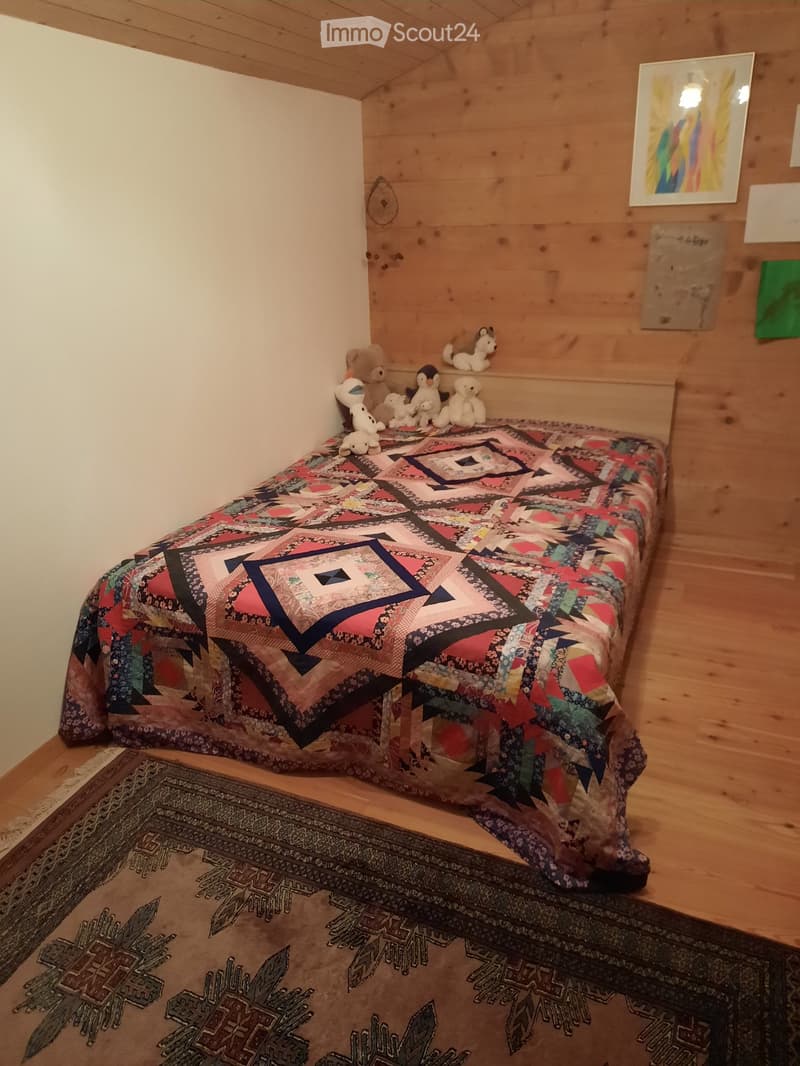 A bedroom with a wooden wall and wooden floor. There is a bed with a colorful quilt, a rug, and several stuffed toys. The room is furnished with an artwork hanging on the wall. The ceiling is wooden.