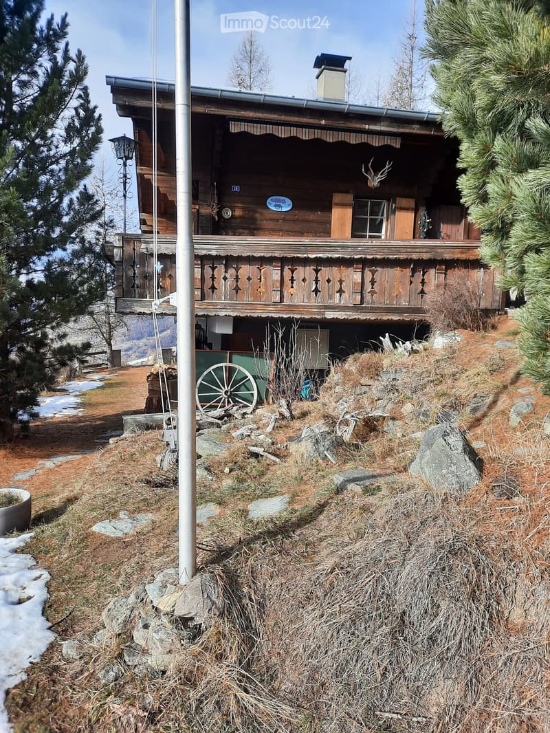 Alpine wooden chalet with raised ground floor, wooden balcony, chimney, green wagon, trees, dry grass, rocks