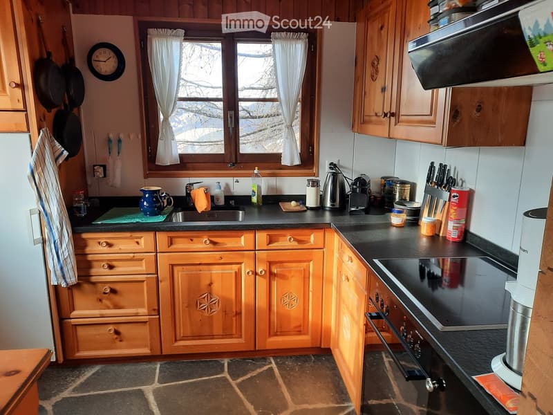 Kitchen with wooden cabinets, granite countertop, gas stove, double sink, window with white curtains, frying pans, clock, wooden floor, stainless steel appliances