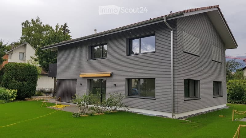 Modern two-story house with grey siding, large windows, and a well-maintained lawn in front.