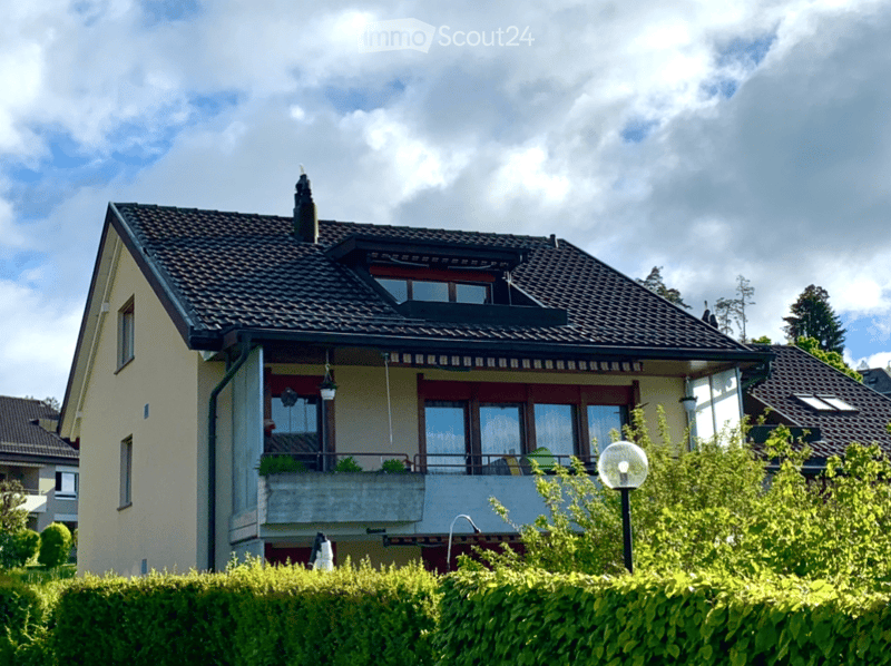 Two-story house with a tiled roof, balcony, and multiple windows, surrounded by greenery.