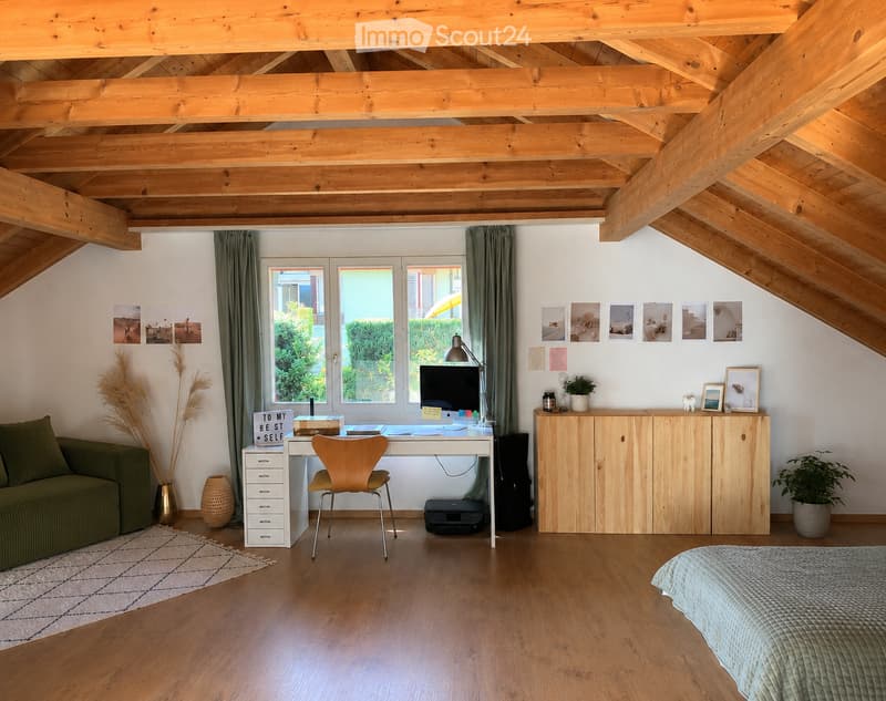 Cozy attic room with exposed wooden beams, featuring a bed, sofa, desk with a chair, and a wooden cabinet. The room has large windows with green curtains, a patterned rug, and decorative plants.