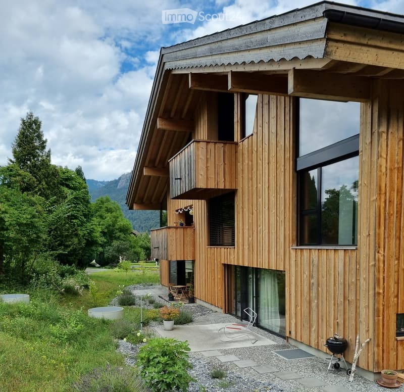 Modern wooden house with large windows, balcony, and outdoor seating area. The house features a contemporary design with a sloped roof and is surrounded by a well-maintained garden and lawn.