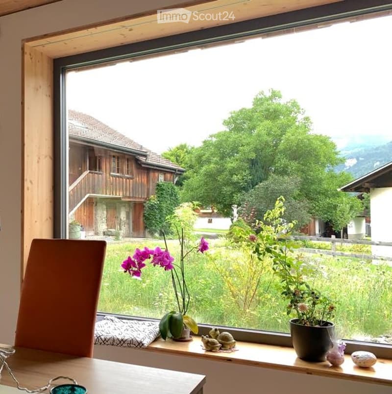 Bright room with large window showcasing a scenic view of a countryside landscape, featuring a wooden table, a chair, and decorative items on the windowsill.