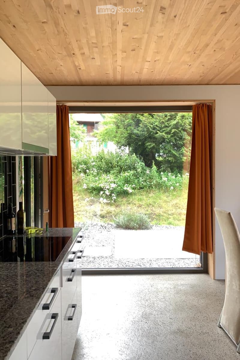 Modern kitchen with white cabinets, granite countertop, large window with brown curtains, and a view of a garden.