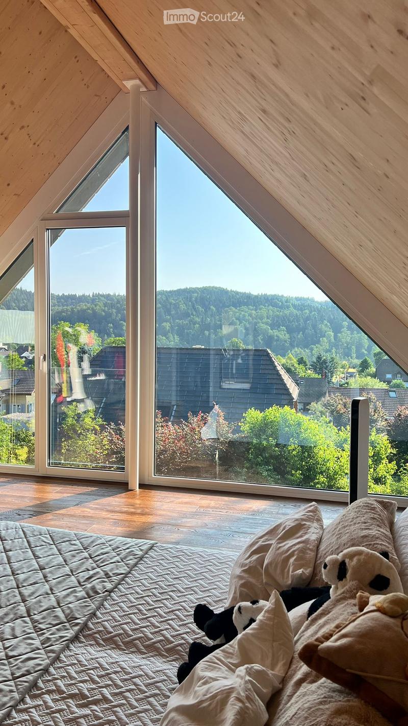 Cozy attic bedroom with wooden slanted ceilings, large windows offering a scenic view, a neatly made bed with pillows and a blanket, and stuffed toys on the bed.